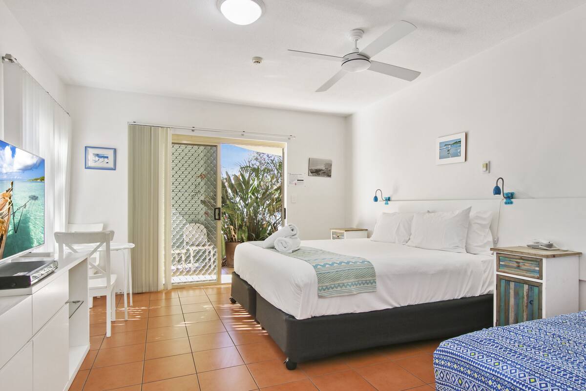 Ein Schlafzimmer mit Bett, Schreibtisch, Stuhl, Deckenventilator und einer Glastür, die auf einen Balkon führt.