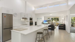A modern kitchen with a large island, stainless steel appliances, and a dining area with a view. - Lennox Beach Resort (Lennox Head)