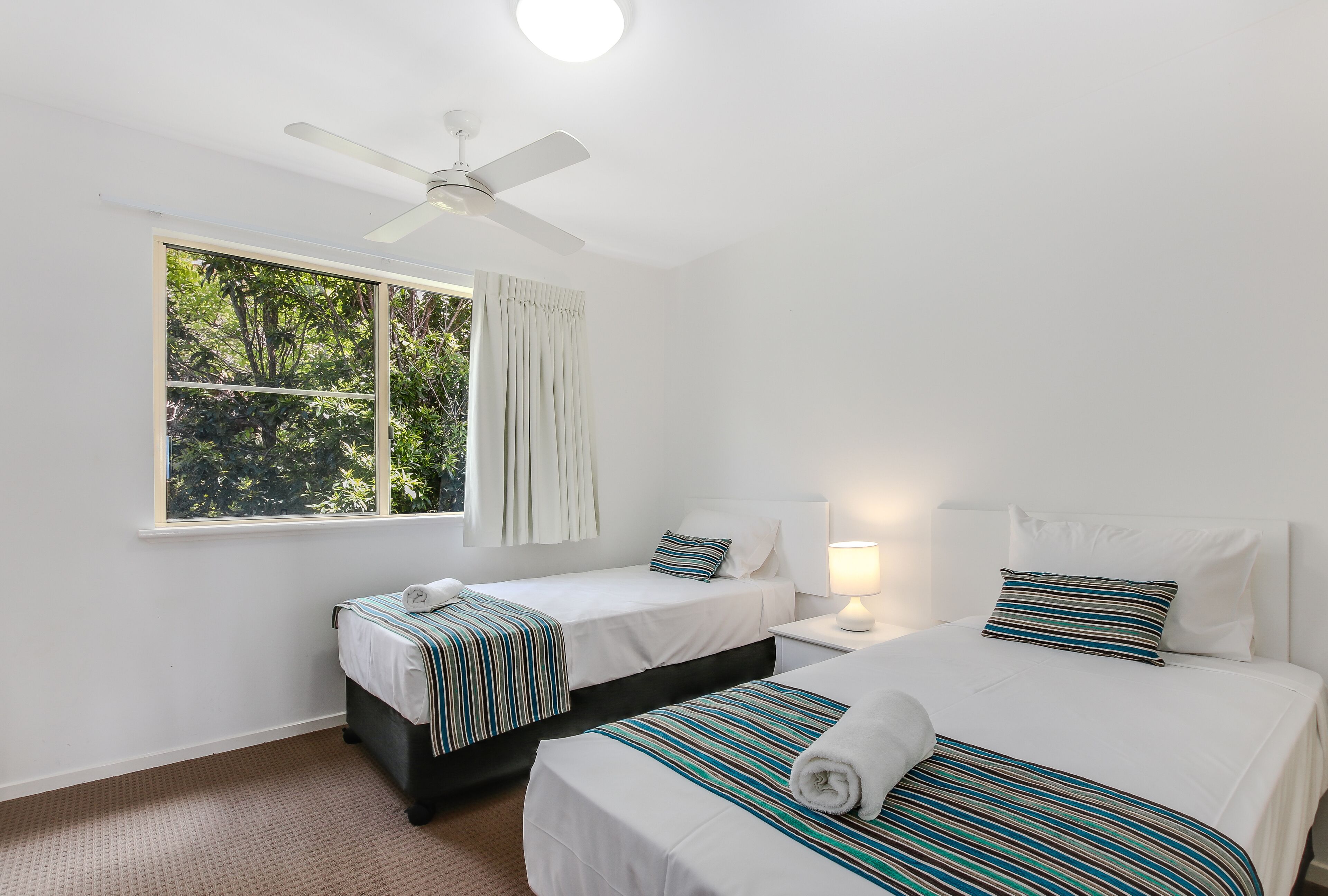 A hotel room with two single beds, a ceiling fan, a window with curtains, and a bedside lamp.