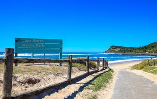 Señal de playa del Parque Marino de Cape Byron, con vista al océano y un sendero de tierra que lleva a la playa.