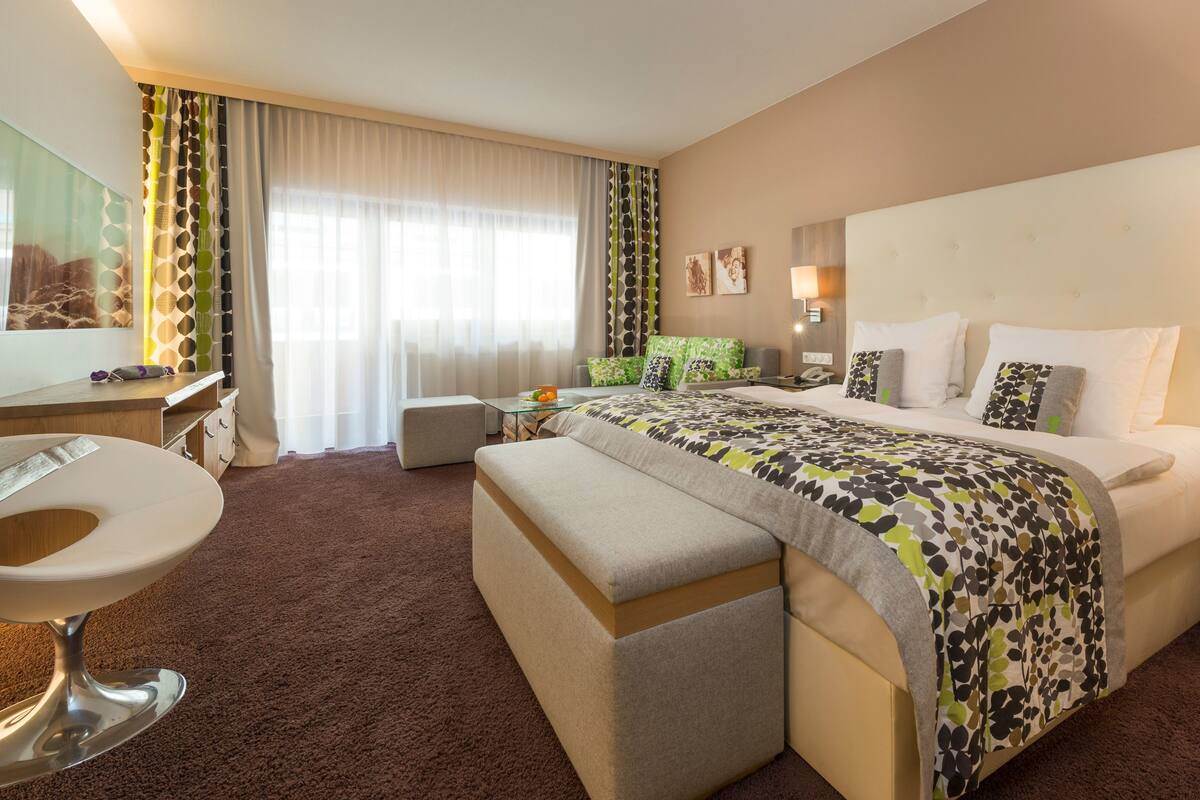 A hotel room with a large bed, a desk, a chair, a small table, and a bedside lamp.