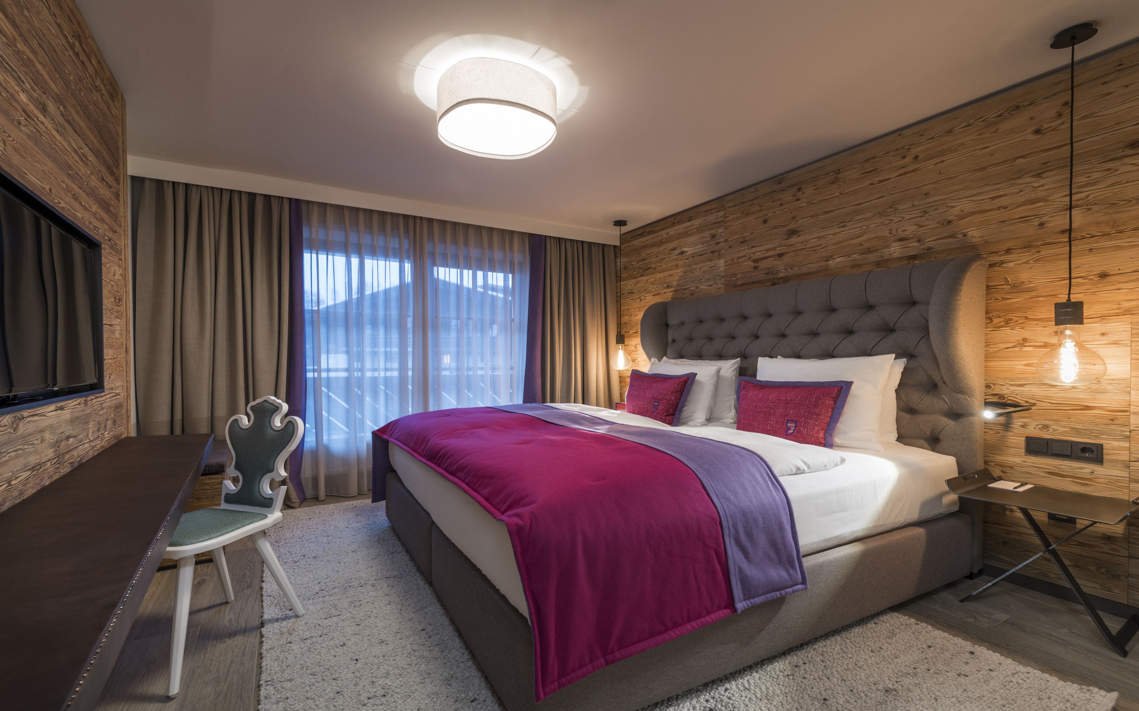 Una camera da letto moderna con una testiera trapuntata, un letto grande con copripiumino viola e bianco, una scrivania con sedia e un'ampia finestra con tende.