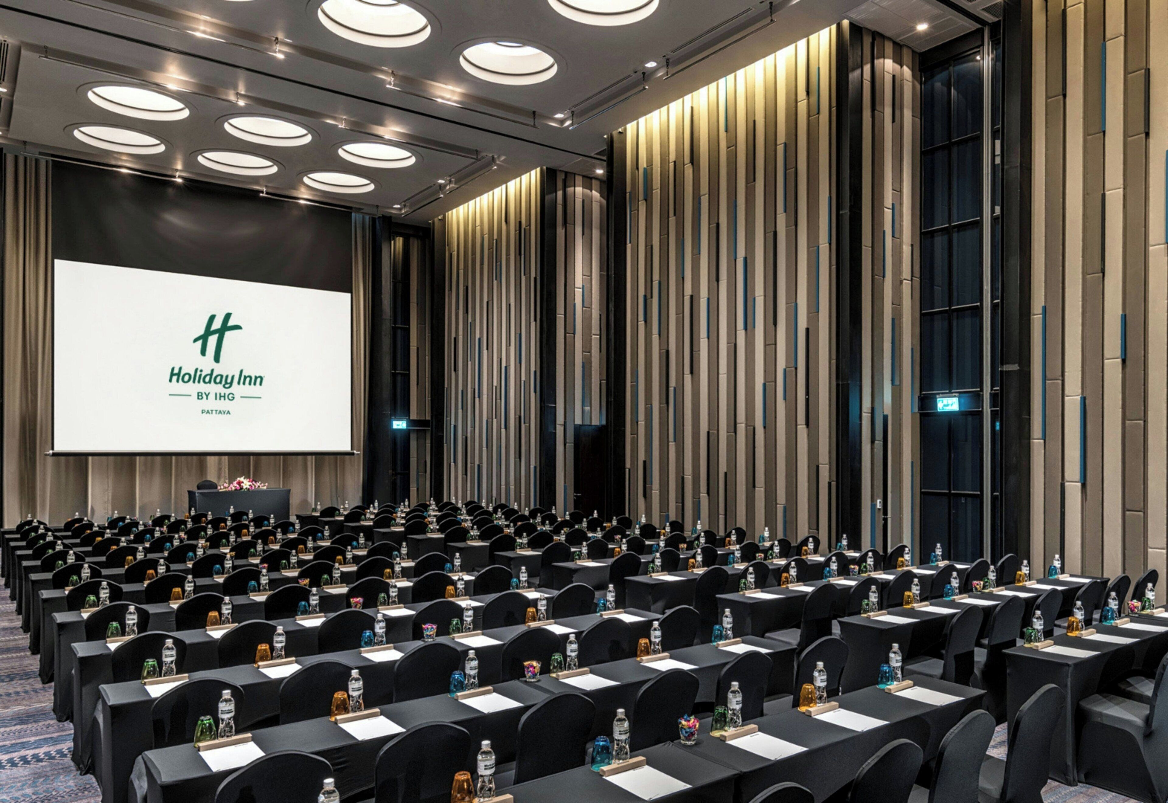 A conference room with rows of tables and chairs, a large screen displaying a presentation, and a modern interior design.