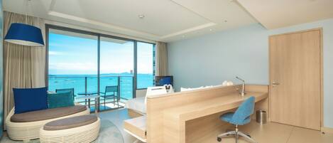 A modern hotel room with a large balcony, a wooden desk, a comfortable seating area, and a view of the sea.