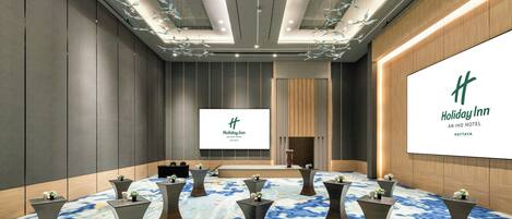 Une salle de conférence dotée d’un grand écran affichant le logo de l’hôtel Holiday Inn Pattaya, de plusieurs tables ornées de centres de table et d’un tapis à motifs.