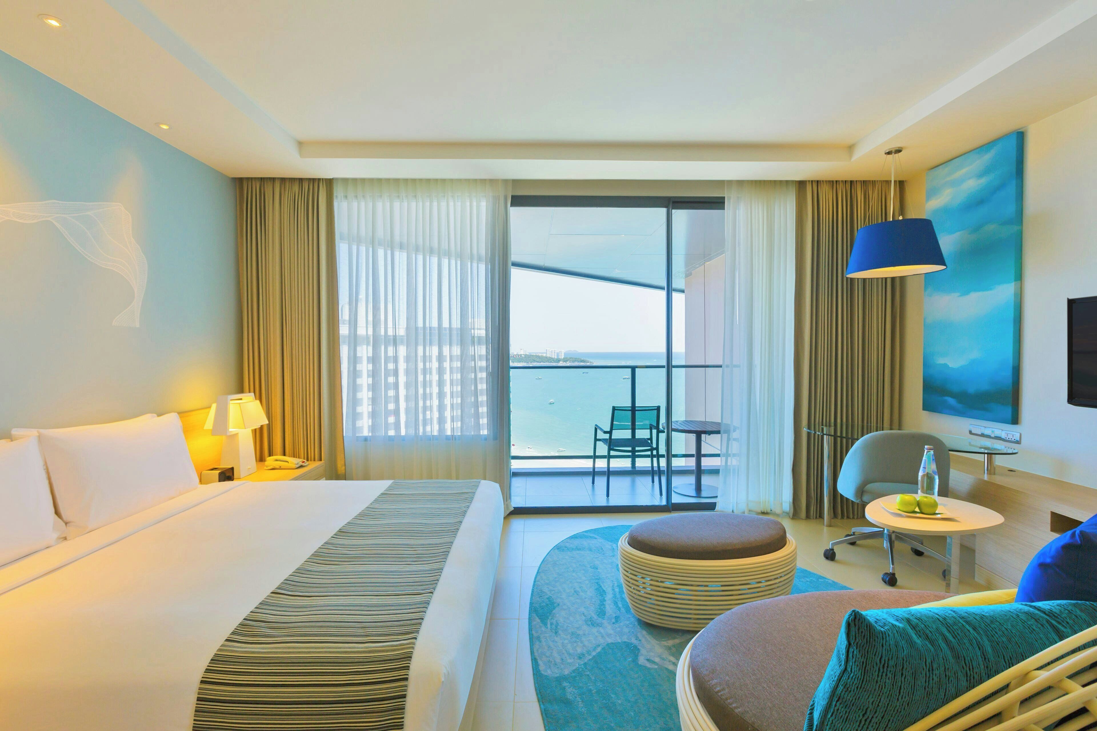 A hotel room with a bed, a desk, a chair, a small table, a sofa, a balcony with a view, and a painting on the wall.