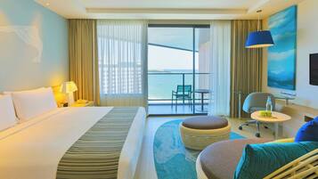 A hotel room with a bed, a desk, a chair, a small table, a sofa, a balcony with a view, and a painting on the wall.