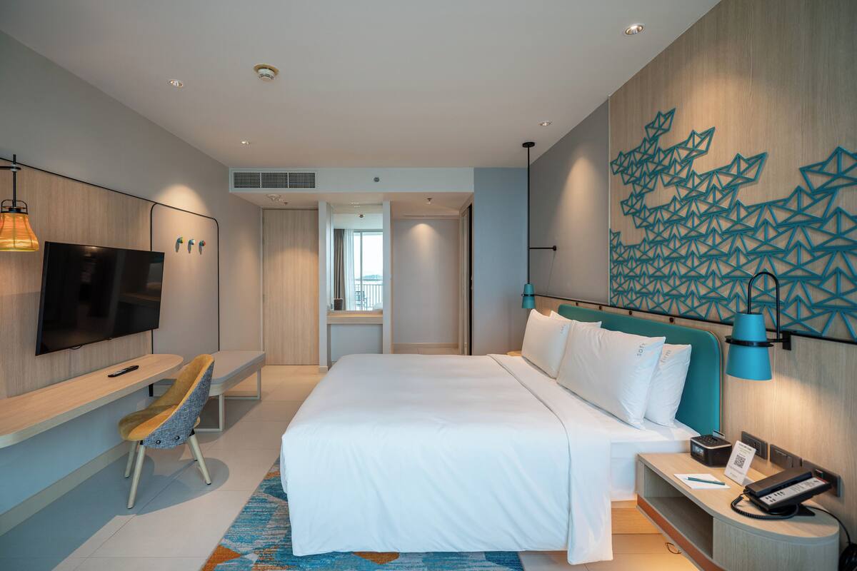 A modern hotel room with a large bed, a desk with a chair, a flat-screen TV, a wall-mounted lamp, and a decorative wall panel.