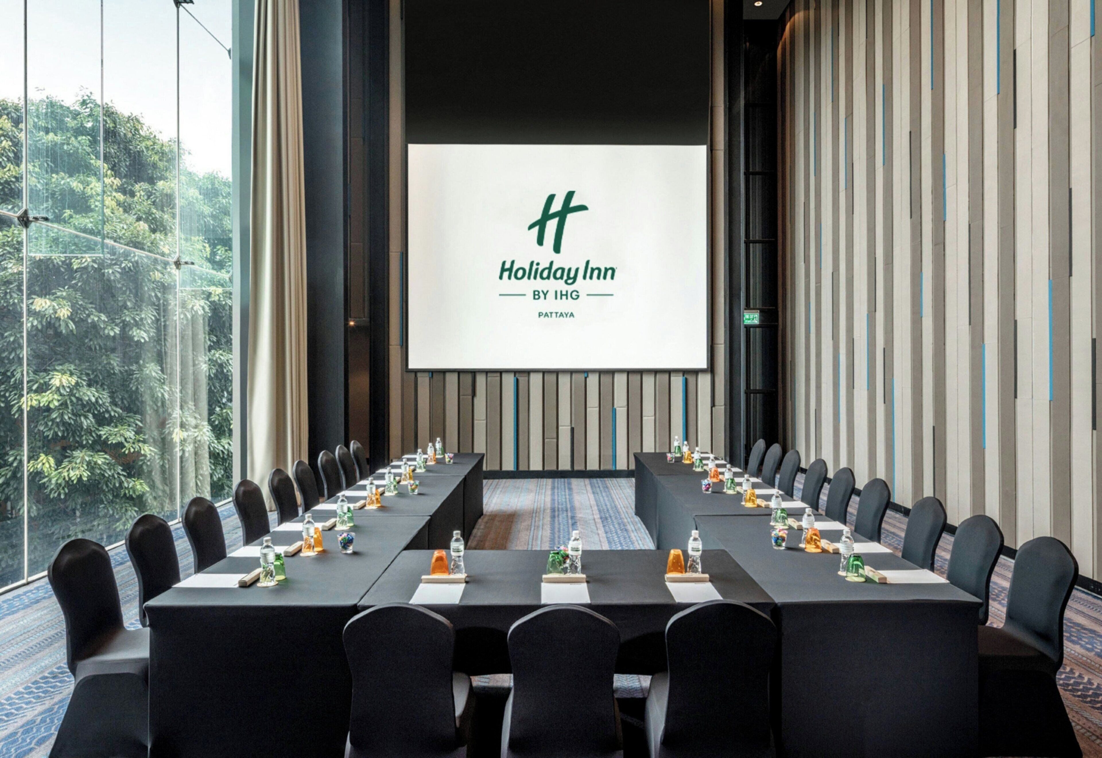 Un salón de conferencias con una larga mesa, sillas y una pantalla que muestra el logotipo de Holiday Inn.
