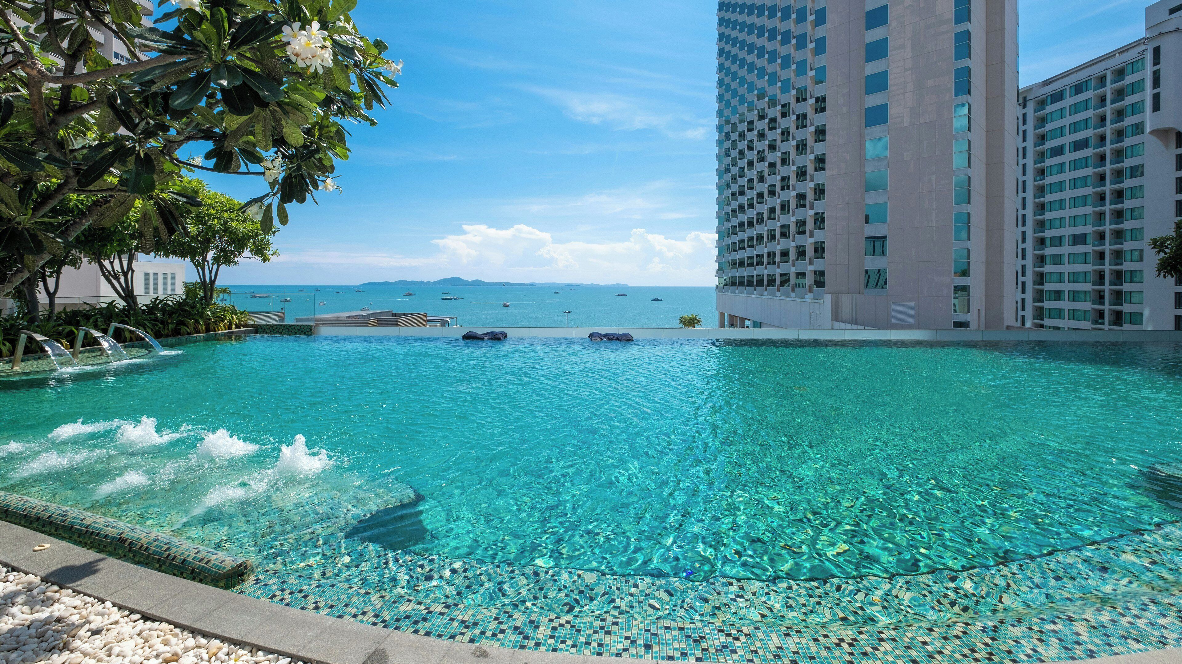 A clear blue pool with a tiled edge, surrounded by a high-rise building and a view of the sea.