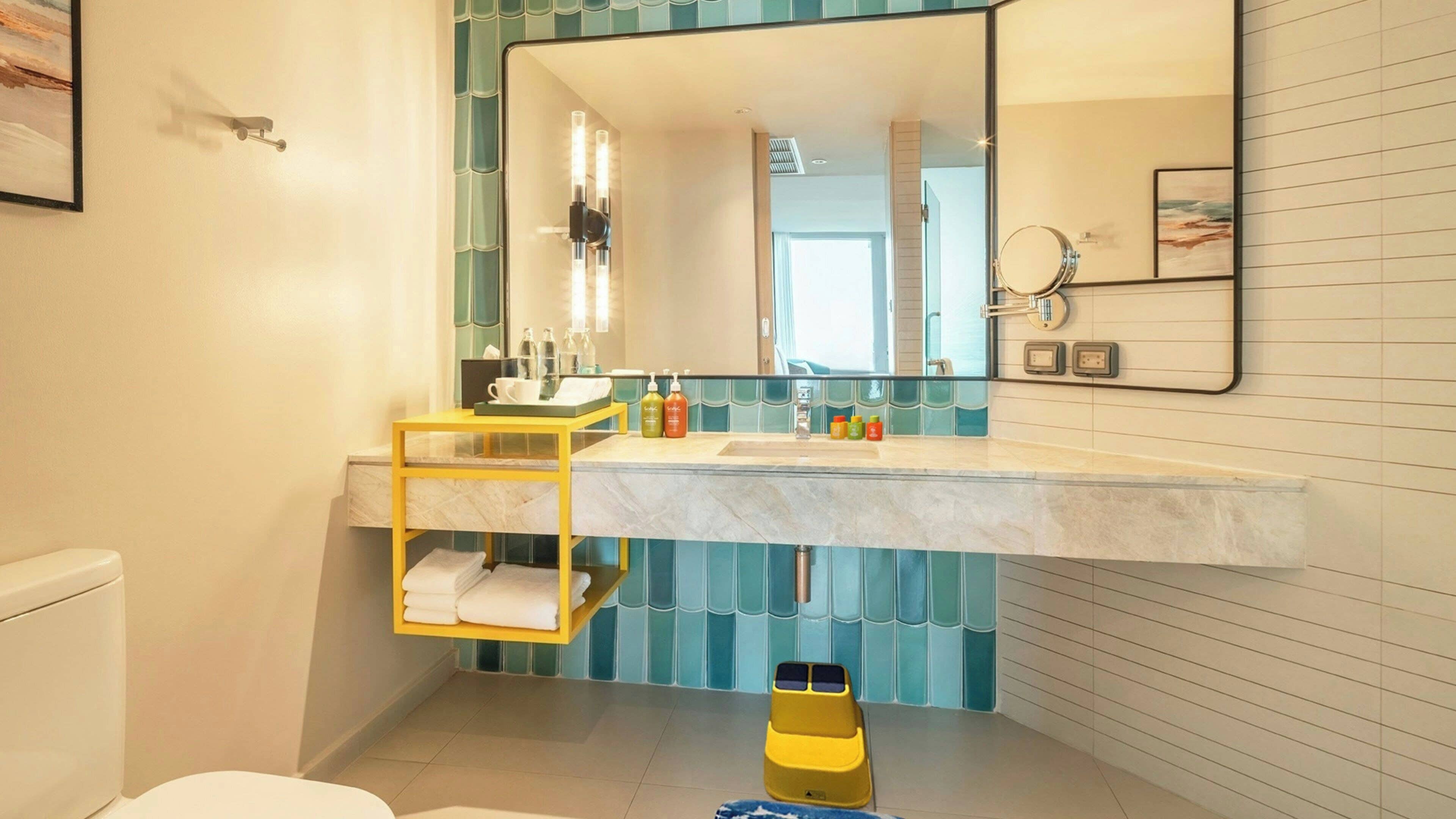 A bathroom with a large mirror, a yellow shelf, a white toilet, and a blue tiled wall.