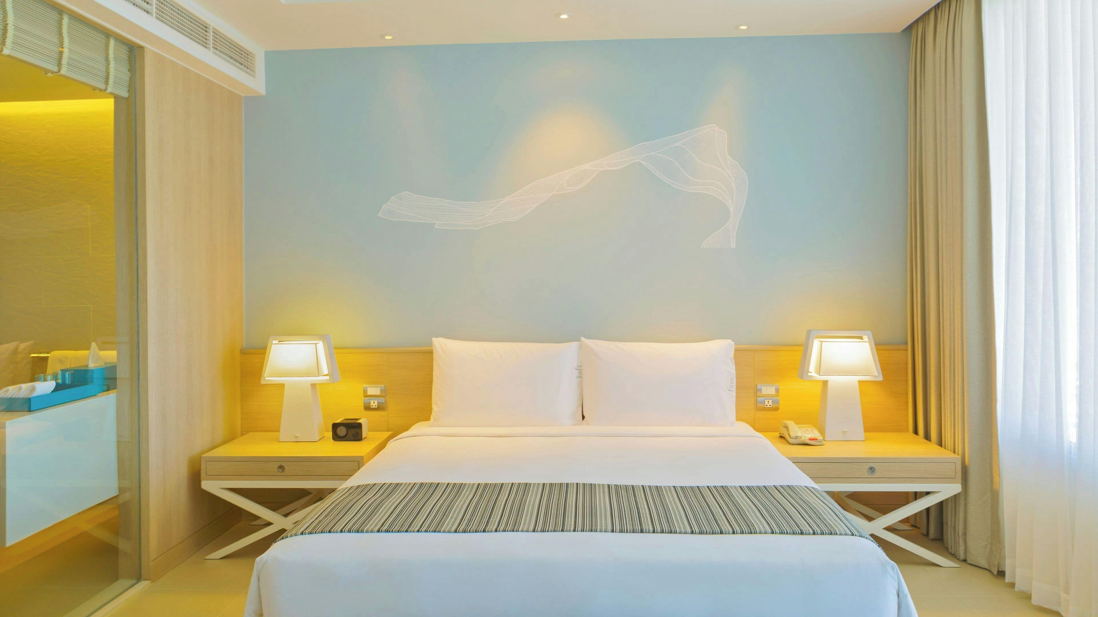 Ein Hotelzimmer mit einem Bett, zwei Nachttischen mit Lampen, einem Wandbild und einem Fenster mit Vorhängen.