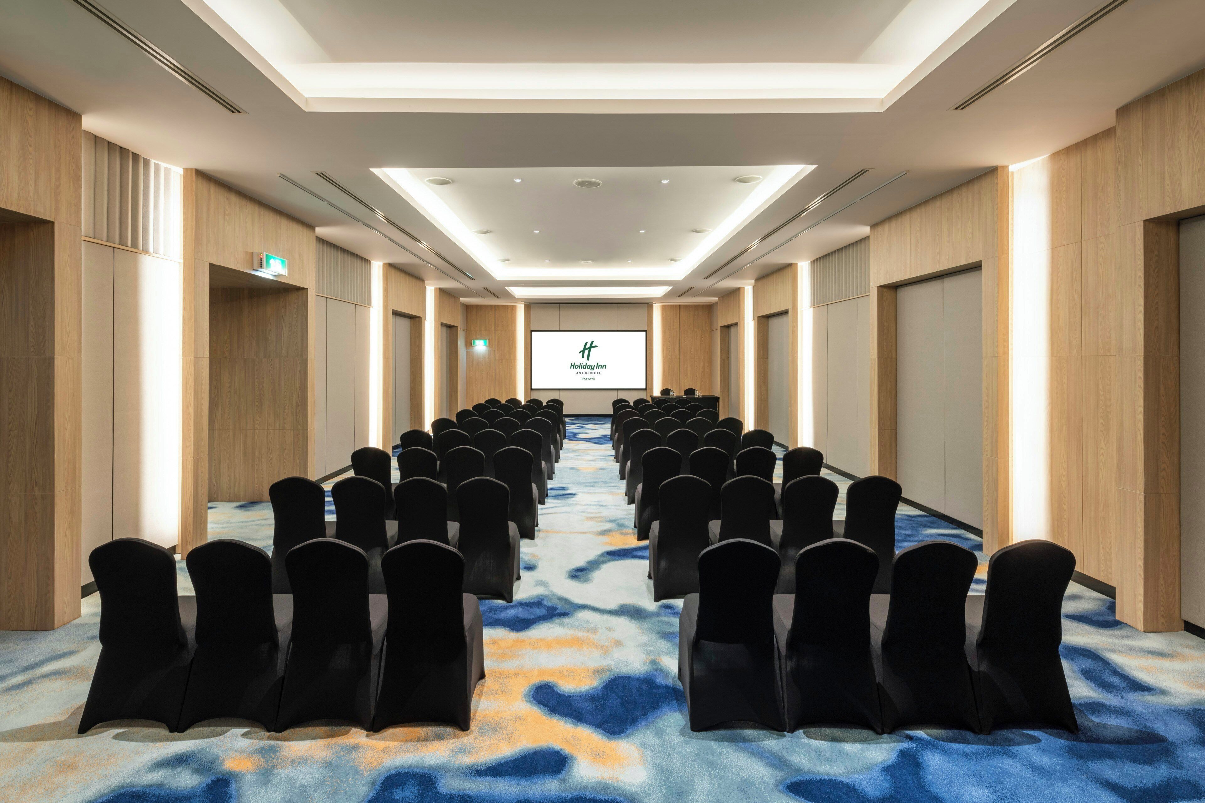 A conference room with rows of chairs facing a screen, wooden panel walls, and a patterned carpet.