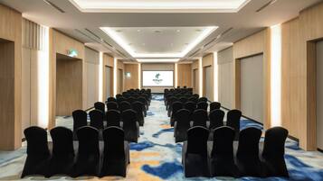 A conference room with rows of chairs facing a screen, wooden panel walls, and a patterned carpet.