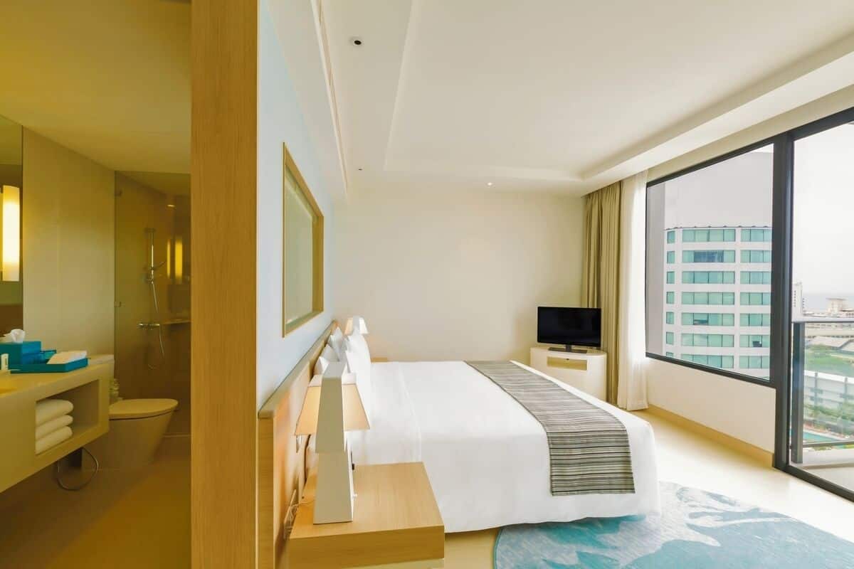 A modern hotel room with a large bed, a TV, a bathroom, and a view of the city.