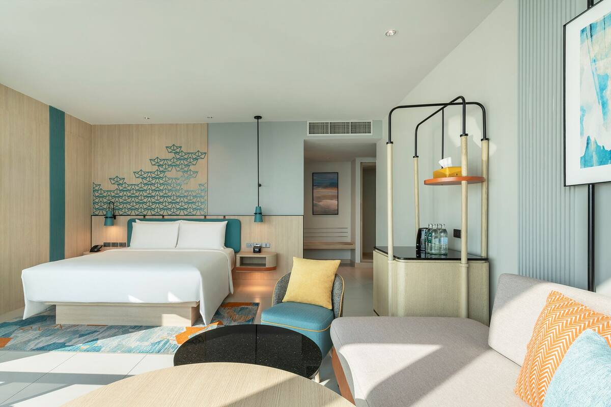 A modern hotel room with a large bed, a sofa, a small table, and a desk with a lamp.
