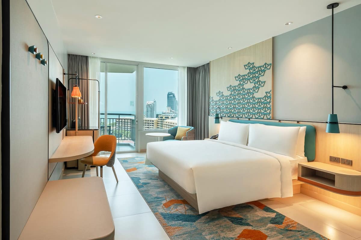 A modern hotel room with a large bed, a desk, and a chair. There is a view of the city through the window.