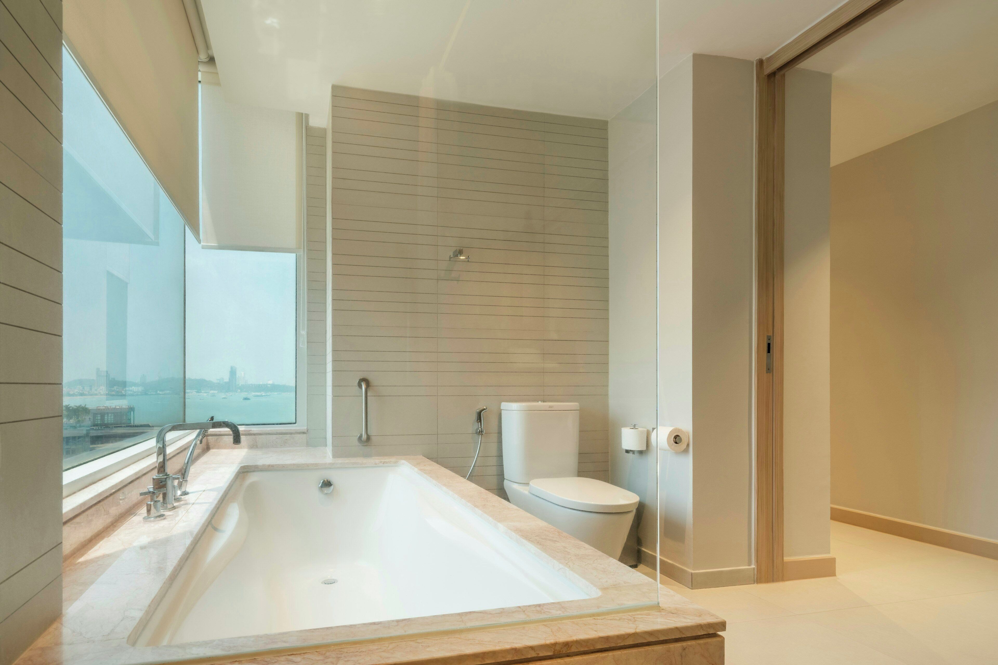 A modern bathroom with a large bathtub, a toilet, and a window offering a city view.