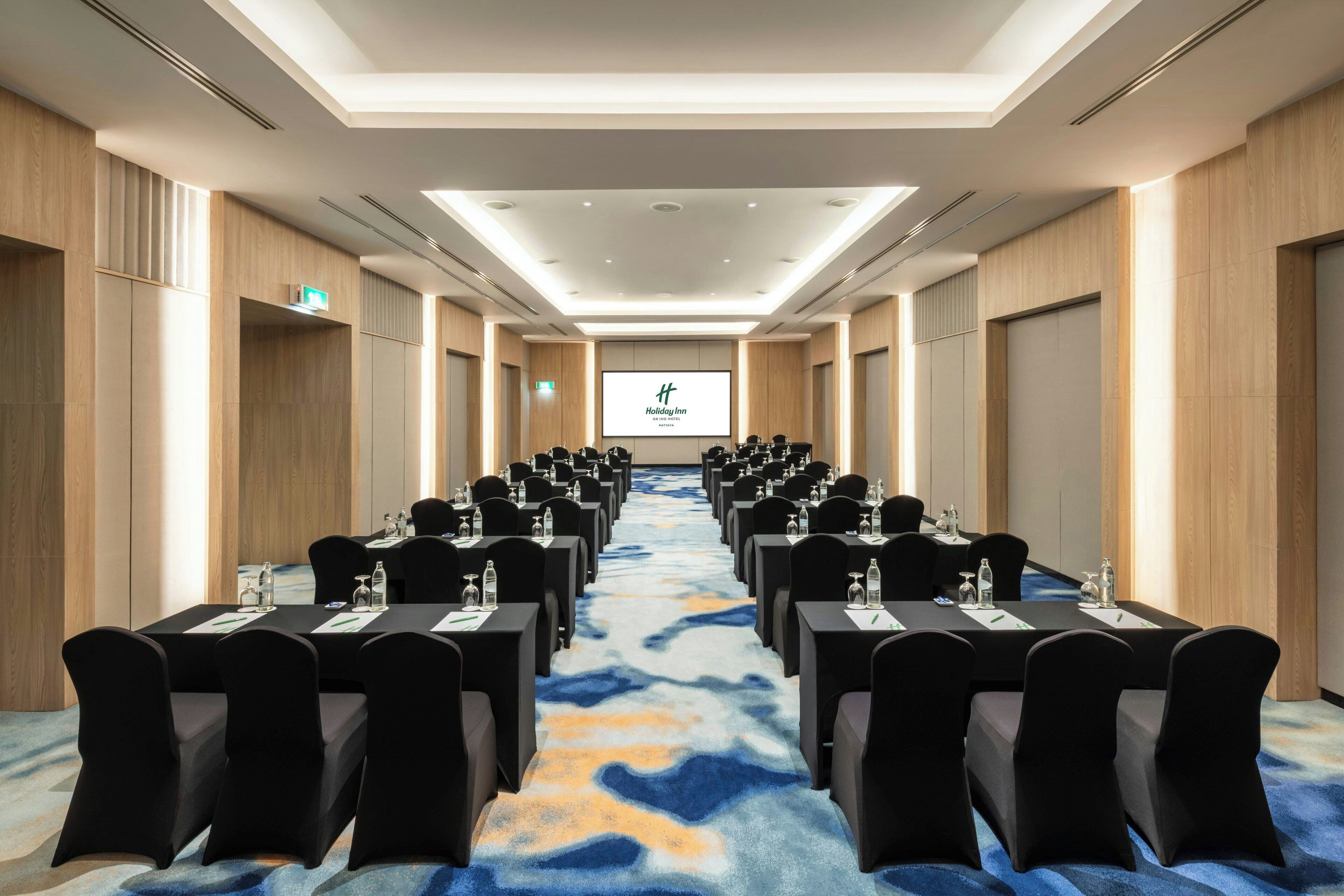Ein Konferenzraum mit Tischen und Stühlen, einer großen Leinwand im vorderen Bereich und einem Teppichboden.