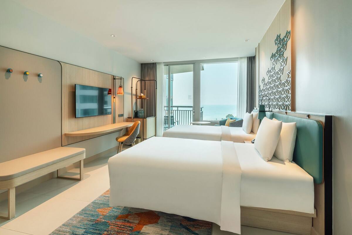 A modern hotel room with a large bed, a desk, and a TV. There is a balcony with a view of the ocean.