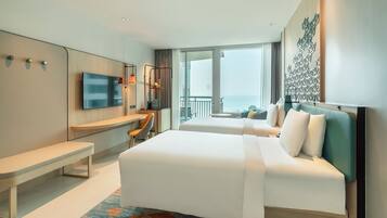 A modern hotel room with a large bed, a desk, and a TV. There is a balcony with a view of the ocean.