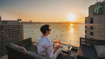 A person seated on a balcony with a view of the sea and a sunset.