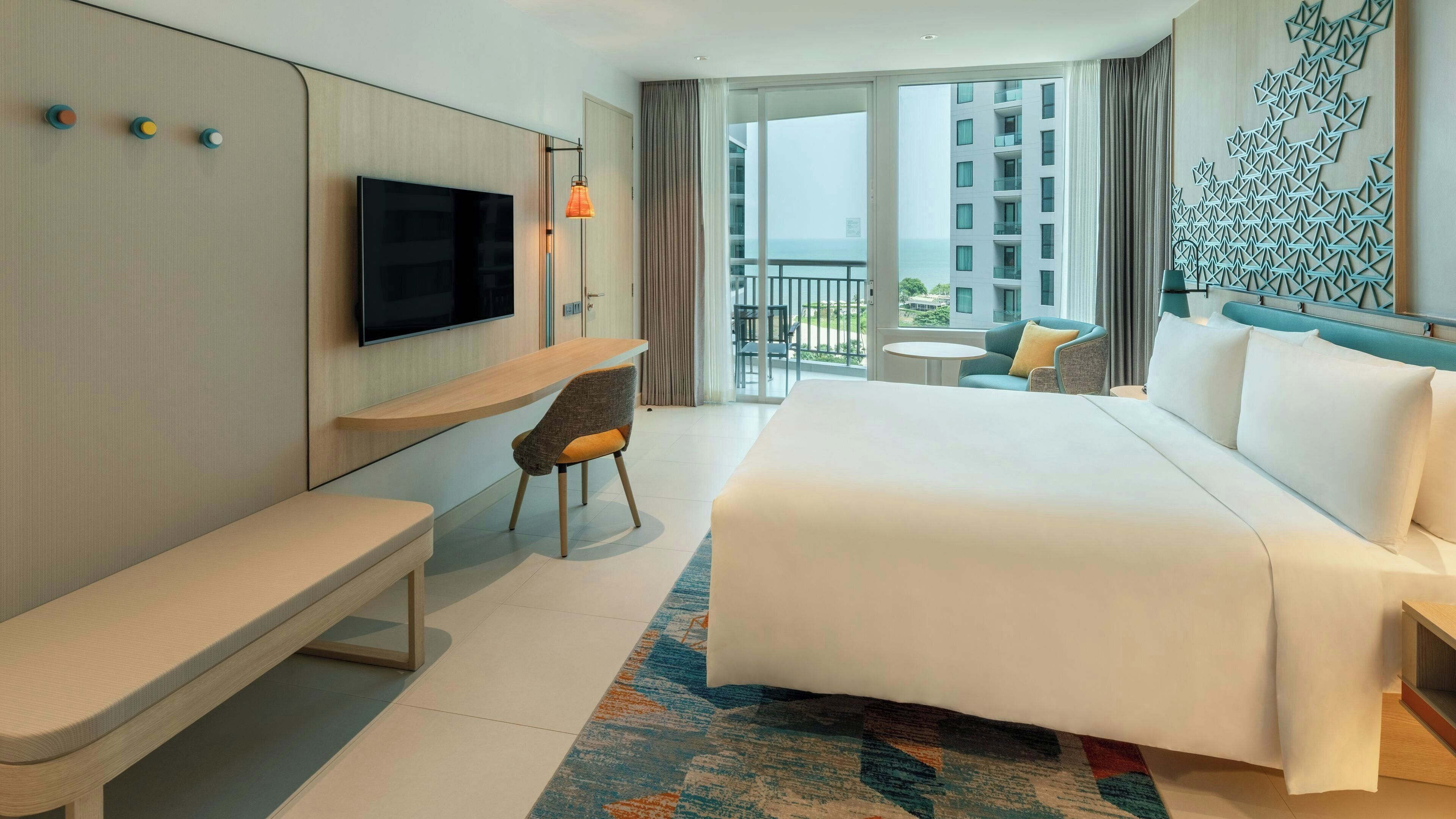 A hotel room with a bed, a desk, a chair, a TV, and a balcony with a view of buildings.
