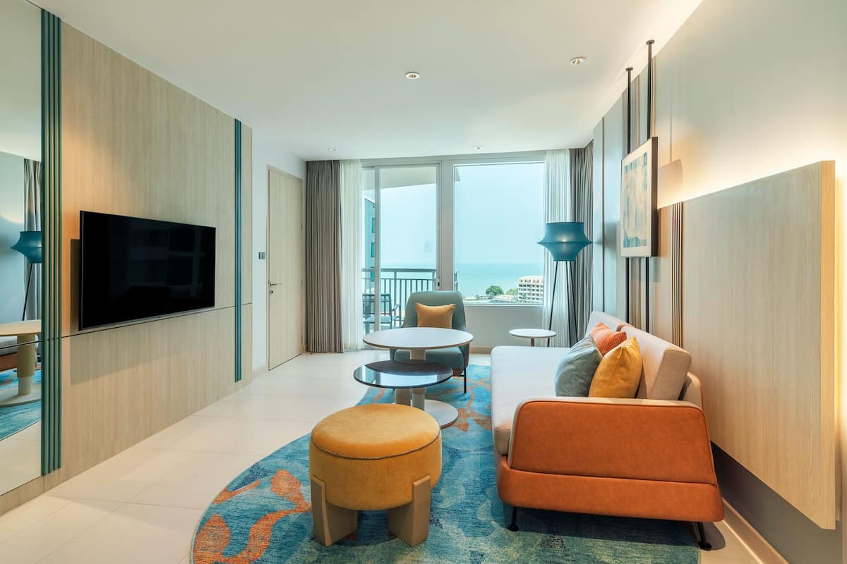 A modern hotel room with a large TV, a sofa, a small table, and a balcony with a view of the sea.