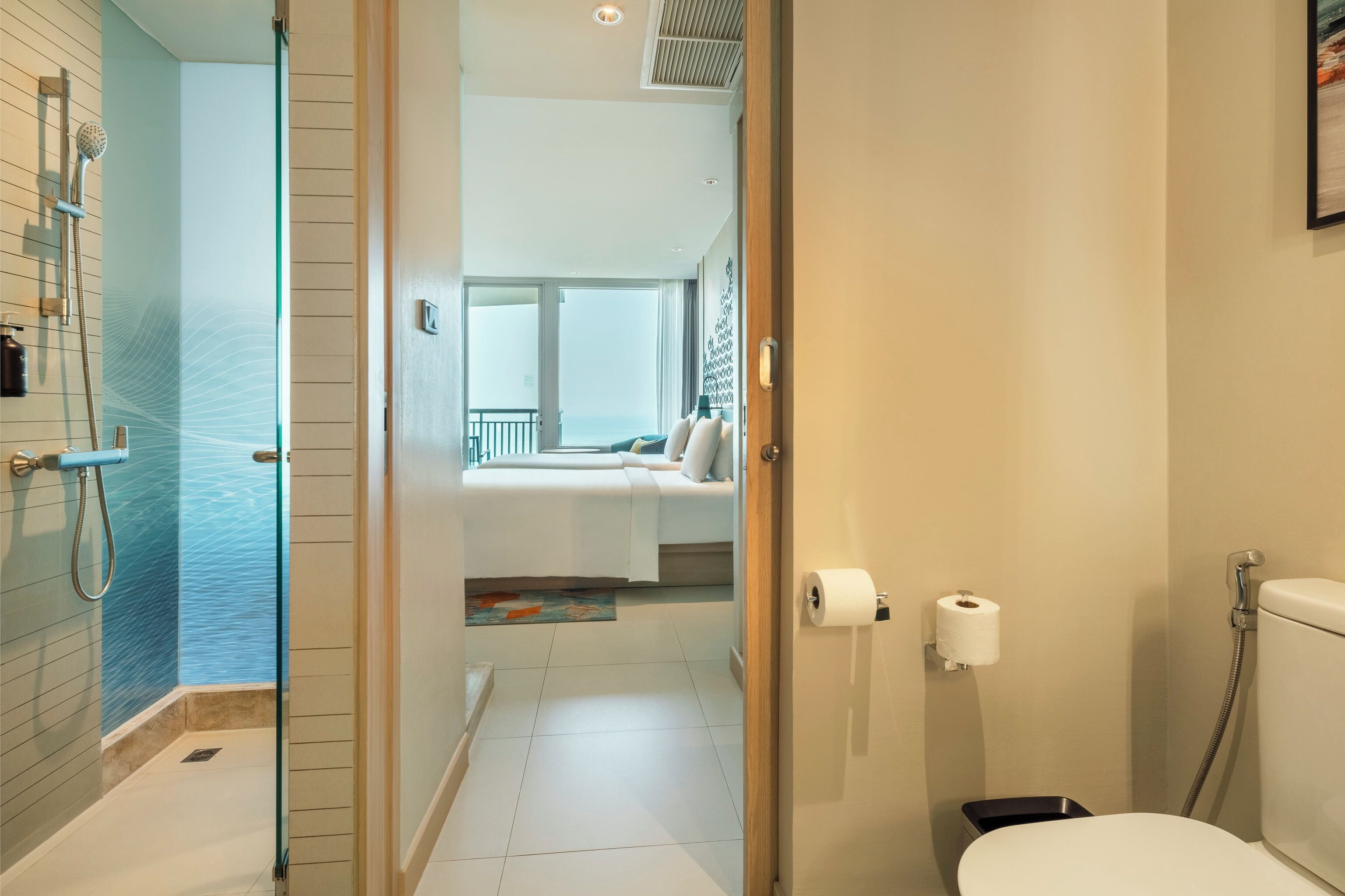 A modern bathroom with a glass shower enclosure, a toilet, and a visible bedroom area with a bed and window.