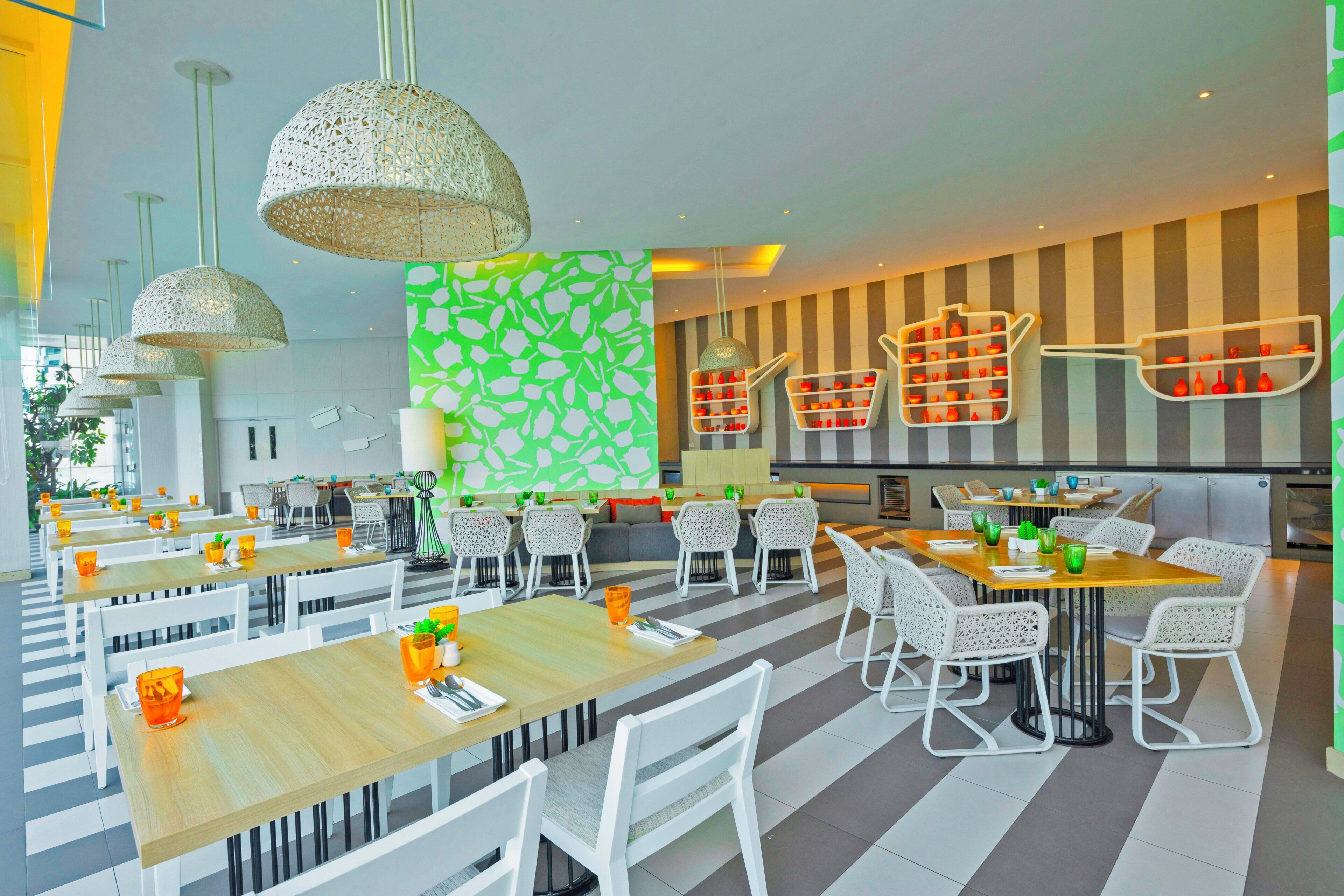 Un interior de restaurante moderno con varias mesas y sillas, lámparas decorativas colgantes y una pared con un estampado verde.