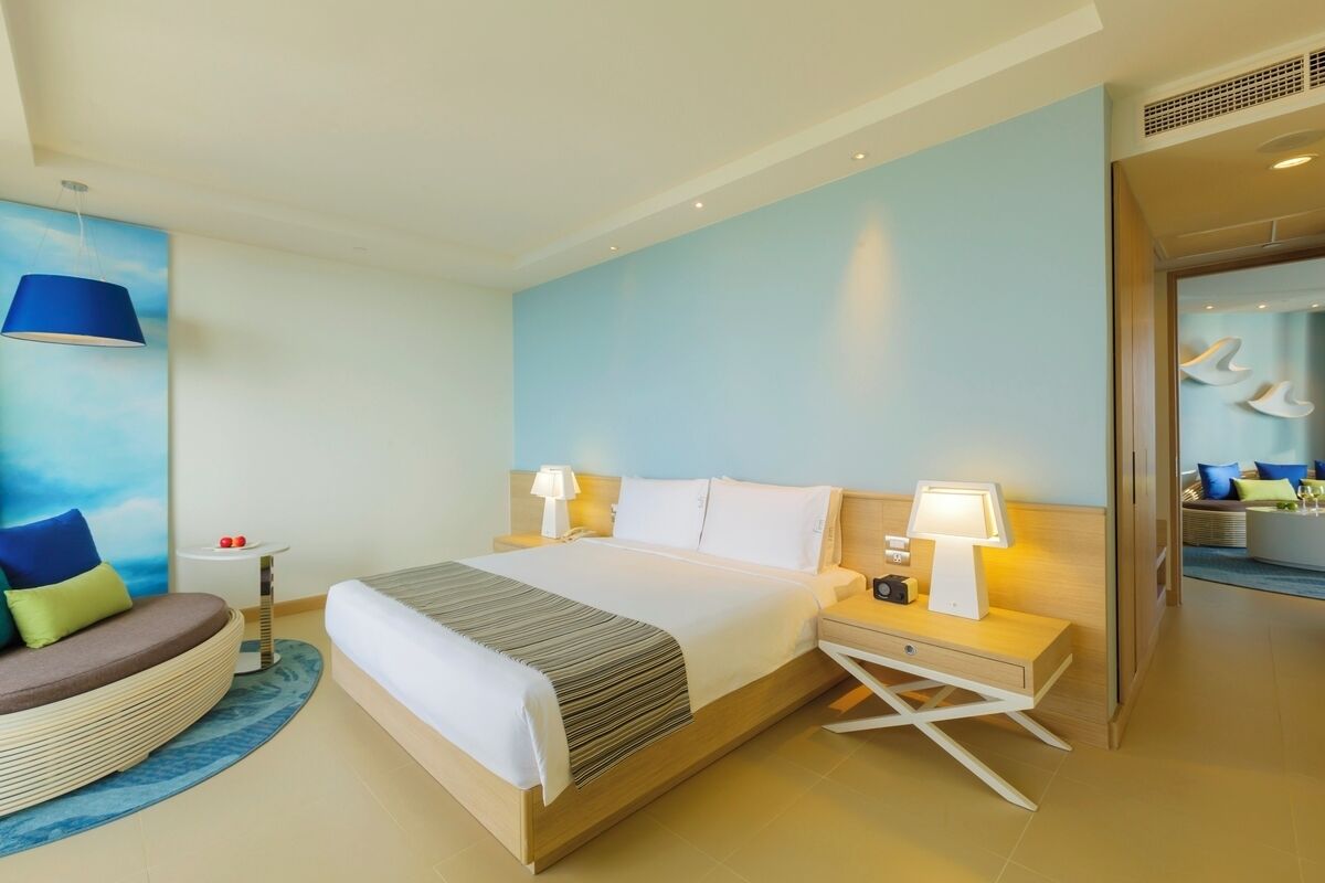 A modern hotel room with a large bed, bedside tables, a sofa, and a view of the sea through the window.