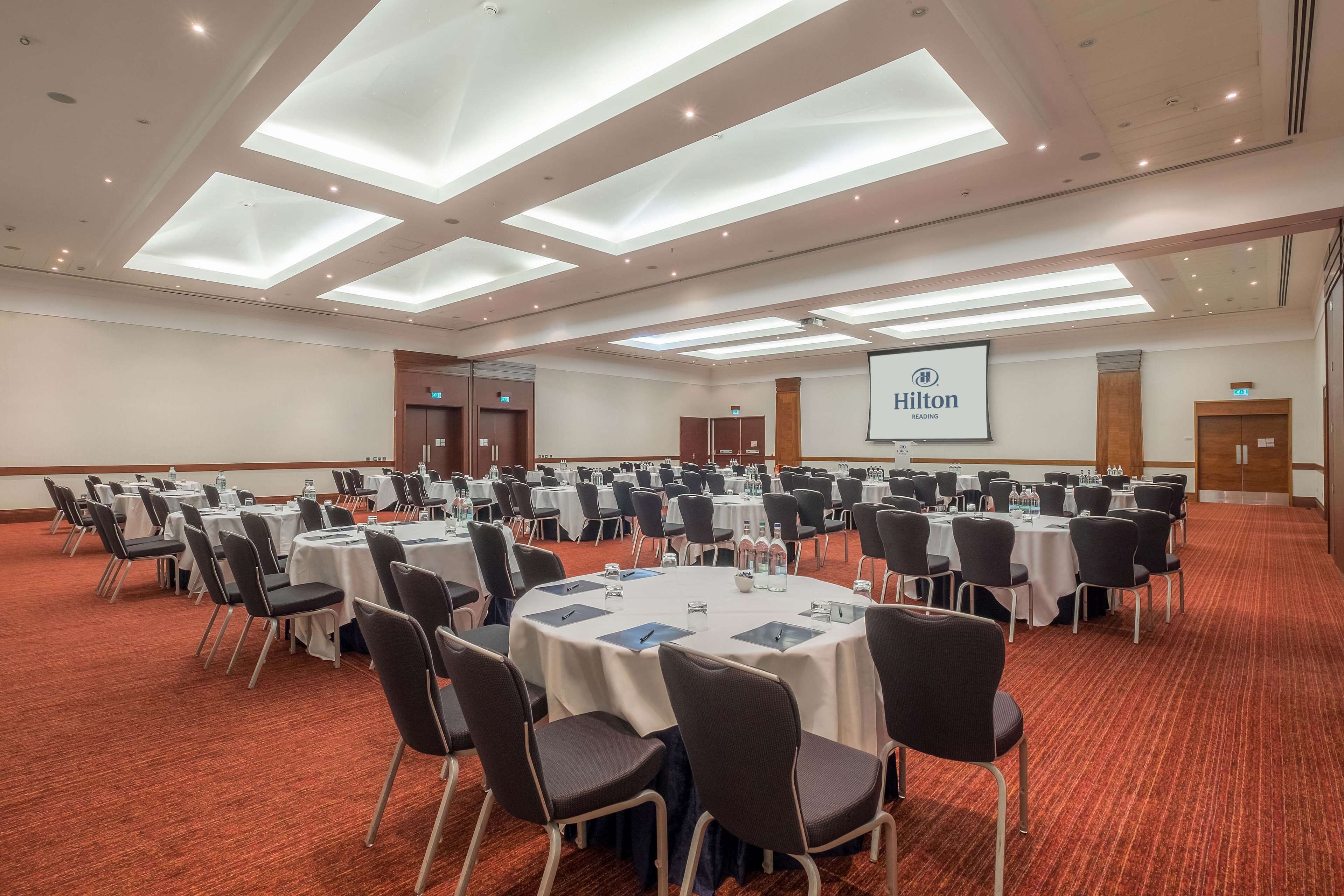 A large, well-lit conference room with multiple round tables and chairs, a projector screen, and a red carpeted floor.