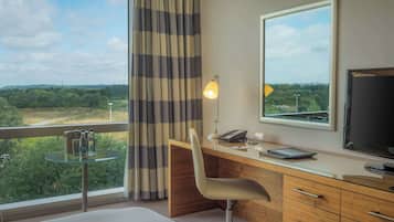 A hotel room with a large window, a desk with a chair, a bed, a small round table with bottles, and a lamp.