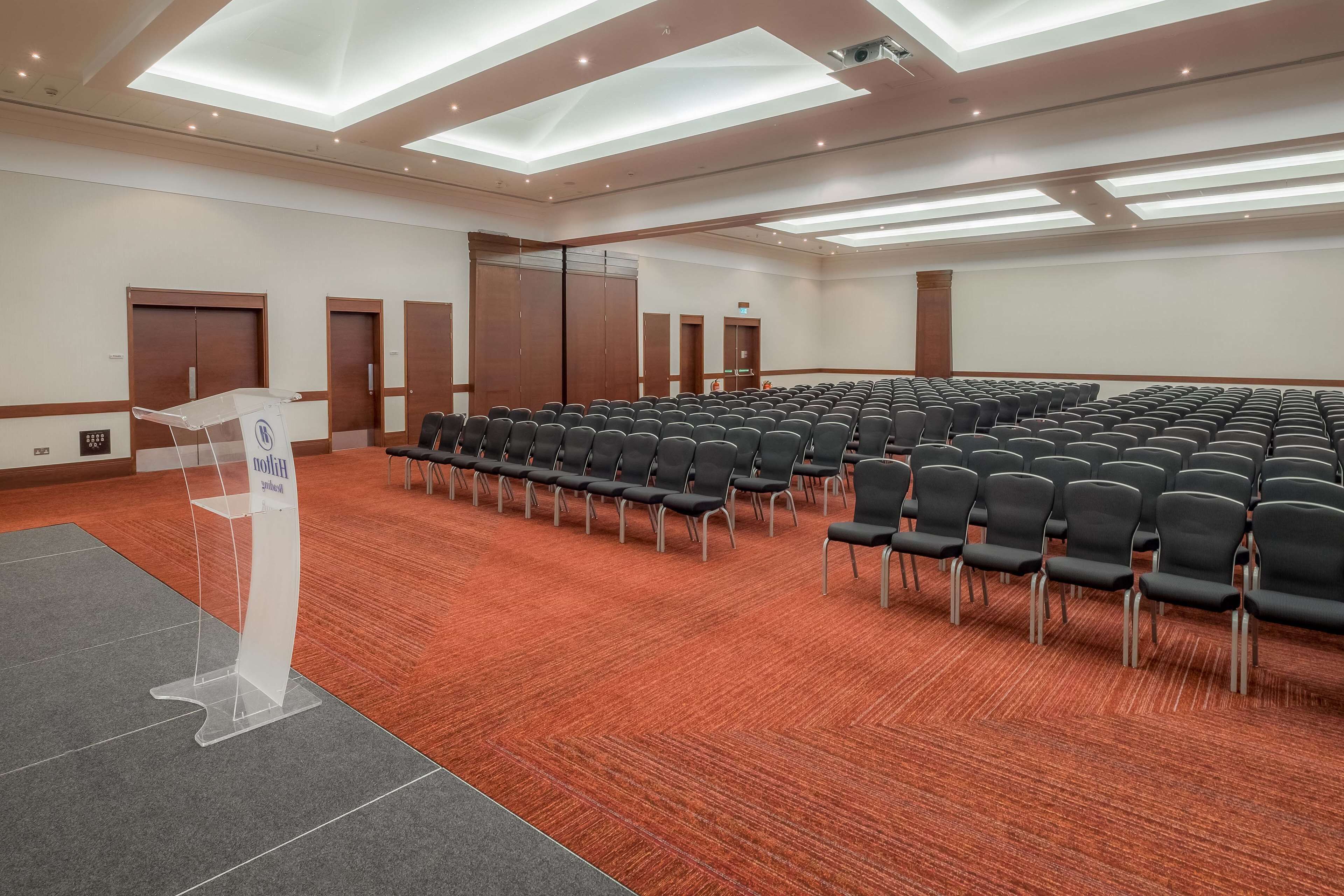 Un salón de conferencias con filas de sillas negras, un atril y puertas de madera.