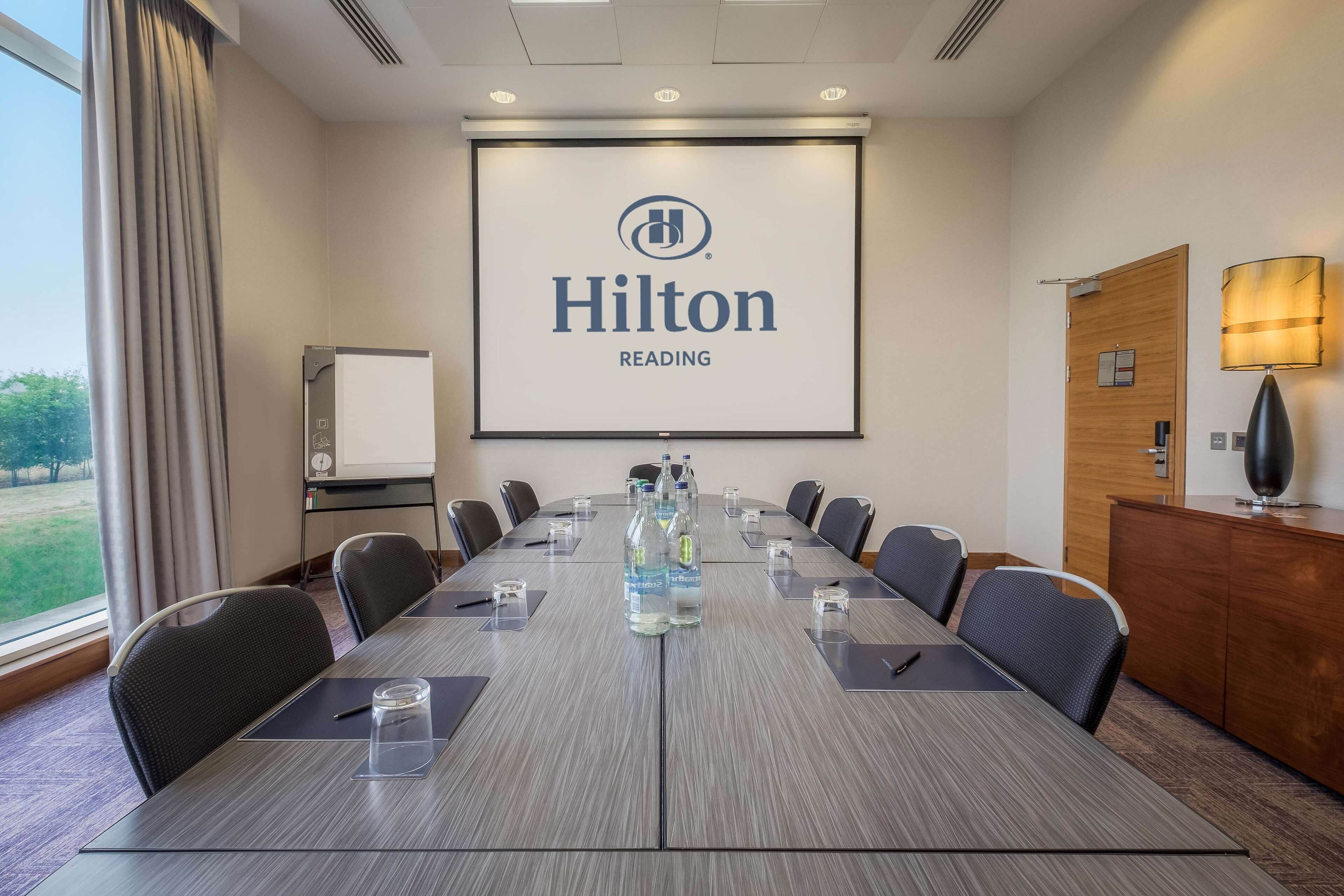 Un salón de conferencias con una gran mesa, sillas, una pantalla de proyector mostrando el logotipo de Hilton Reading, un armario de madera y una lámpara.
