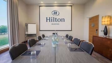 Un salón de conferencias con una gran mesa, sillas, una pantalla de proyector mostrando el logotipo de Hilton Reading, un armario de madera y una lámpara.