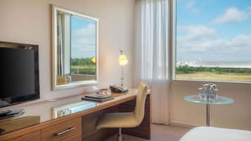 A hotel room with a desk, chair, TV, and a view of a building through the window.