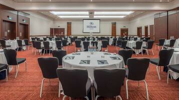 A large conference room with round tables, black chairs, and a projector screen displaying the Hilton logo.