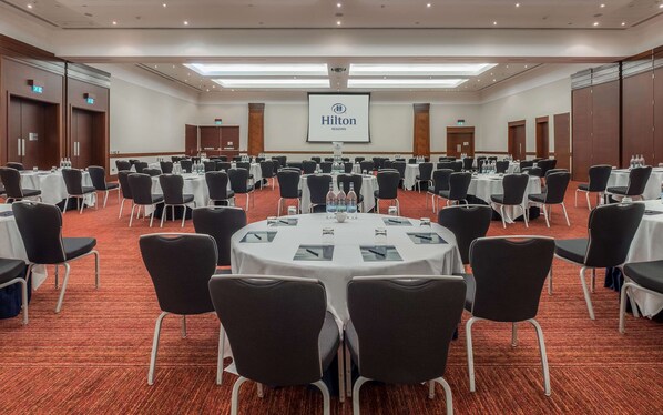 Un amplio salón de conferencias con mesas redondas, sillas negras y una pantalla de proyector que muestra el logotipo de Hilton.