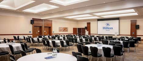 A large conference room with multiple round tables and chairs, a projector screen with the Hilton logo, and a carpeted floor.