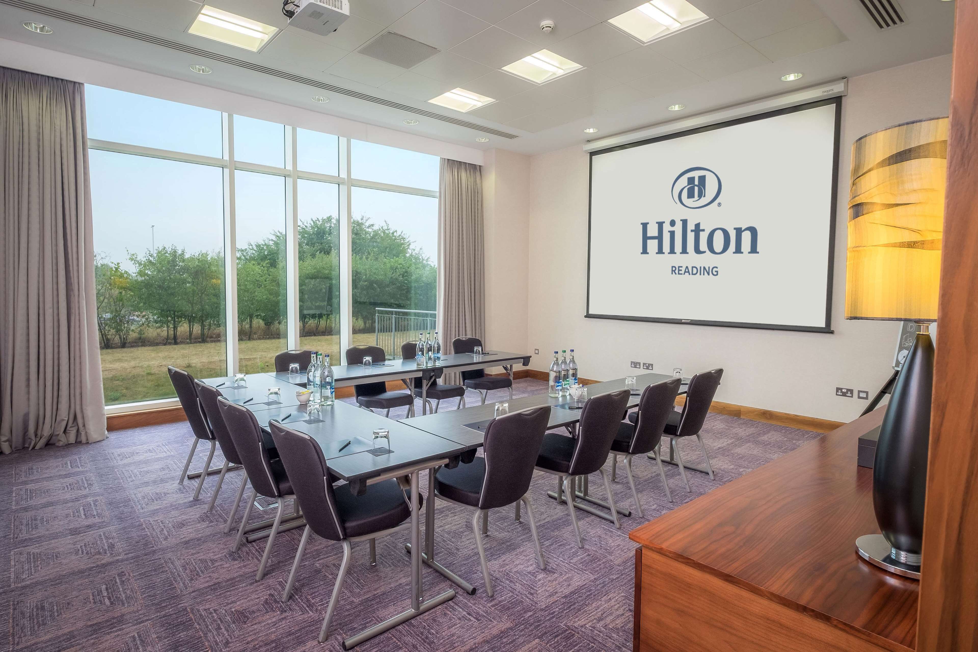 Un salón de conferencias con una gran mesa, sillas, una pantalla de proyector mostrando 'Hilton Reading' y vistas a zonas verdes al exterior.