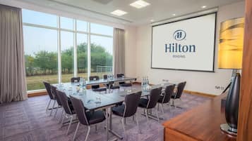 Un salón de conferencias con una gran mesa, sillas, una pantalla de proyector mostrando 'Hilton Reading' y vistas a zonas verdes al exterior.