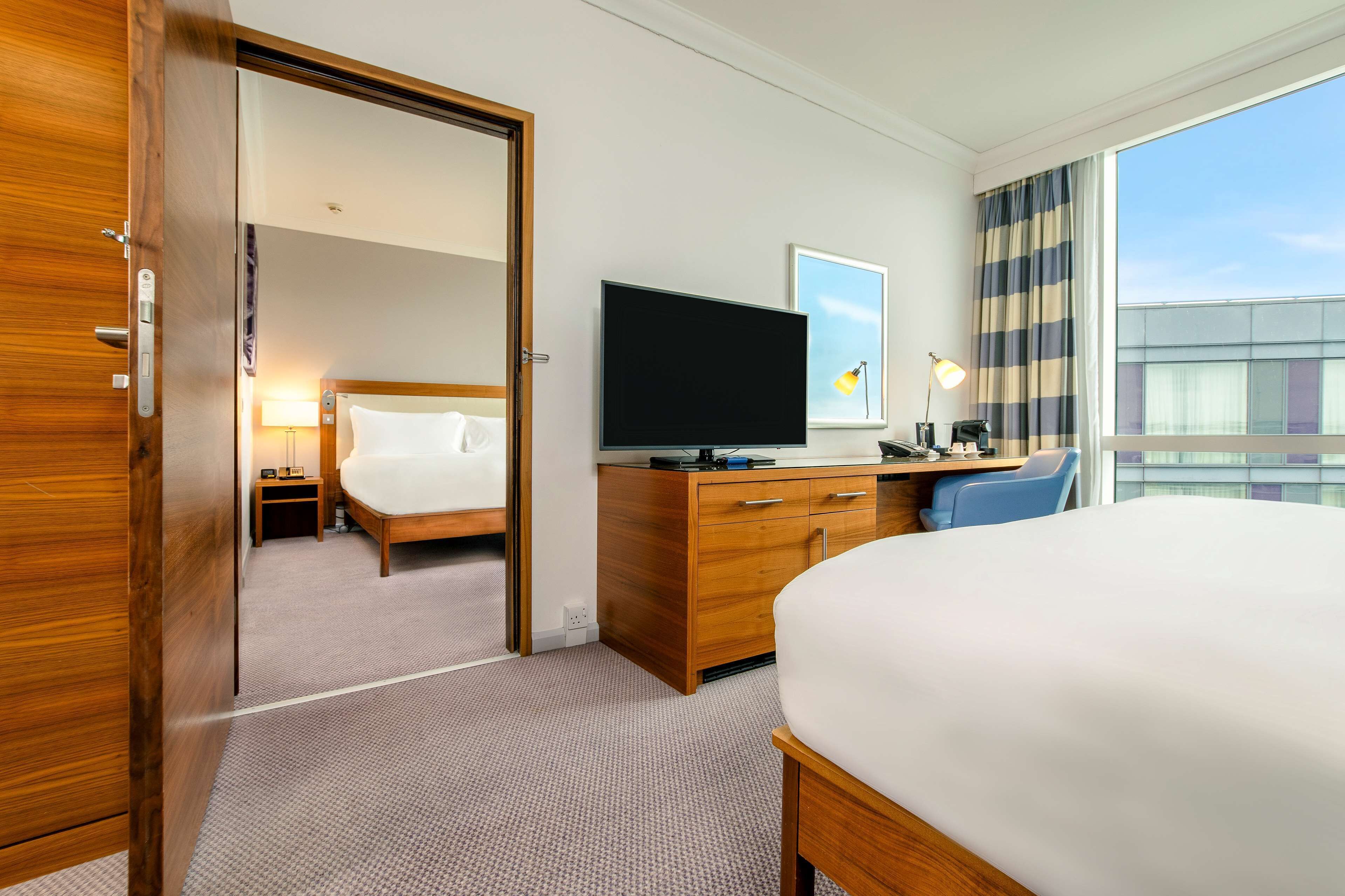 A hotel room with a large bed, a television, a desk with a lamp, and a view of the city through the window.