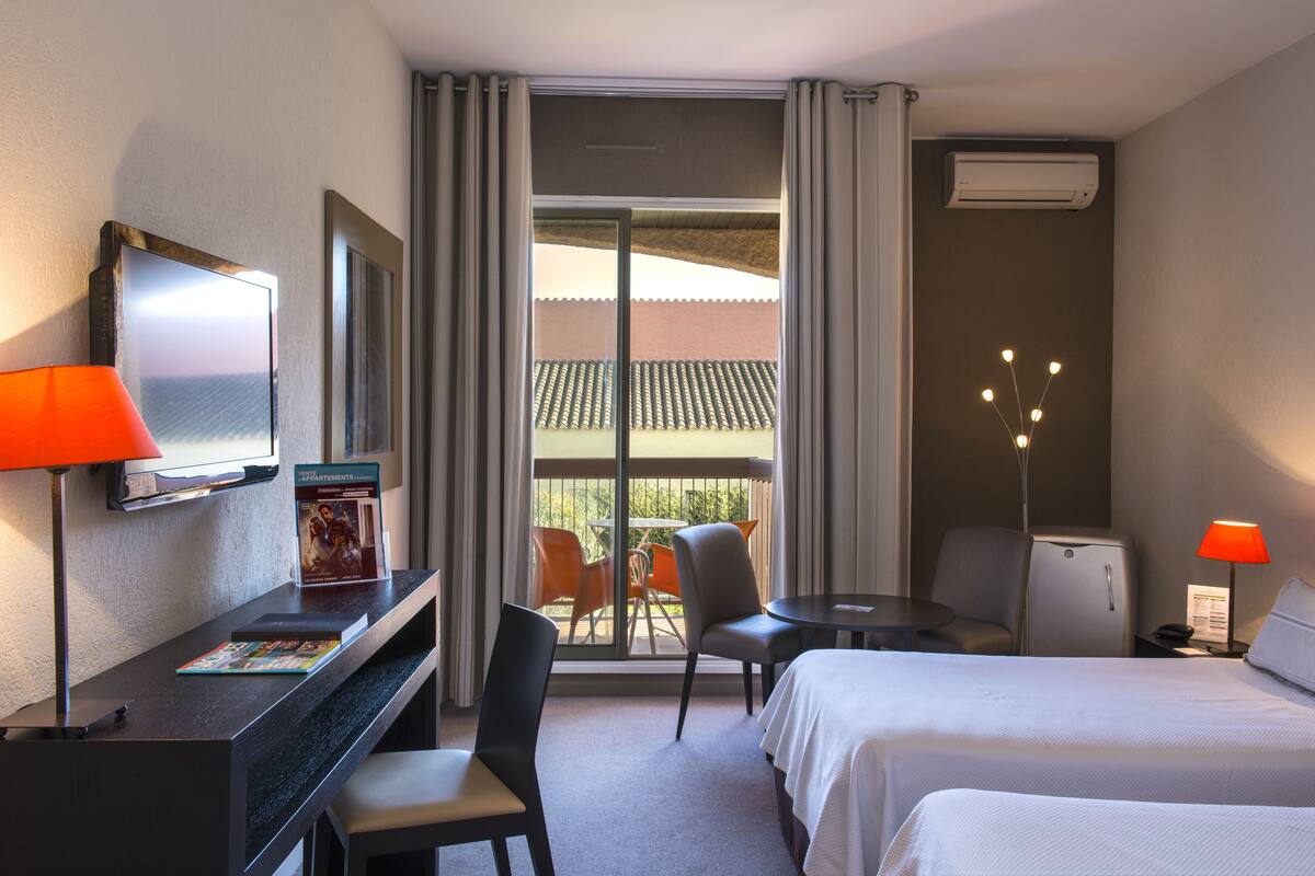 Quarto de hotel com escrivaninha, duas cadeiras, uma cama, uma mesinha e uma janela com cortinas.