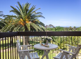 Un balcón con dos sillas blancas y una pequeña mesa redonda, con vistas a un jardín con palmeras y montañas a lo lejos.