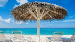 A beach with white lounge chairs and a thatched umbrella. - Aqua Bay Club Condos (Seven Mile Beach)