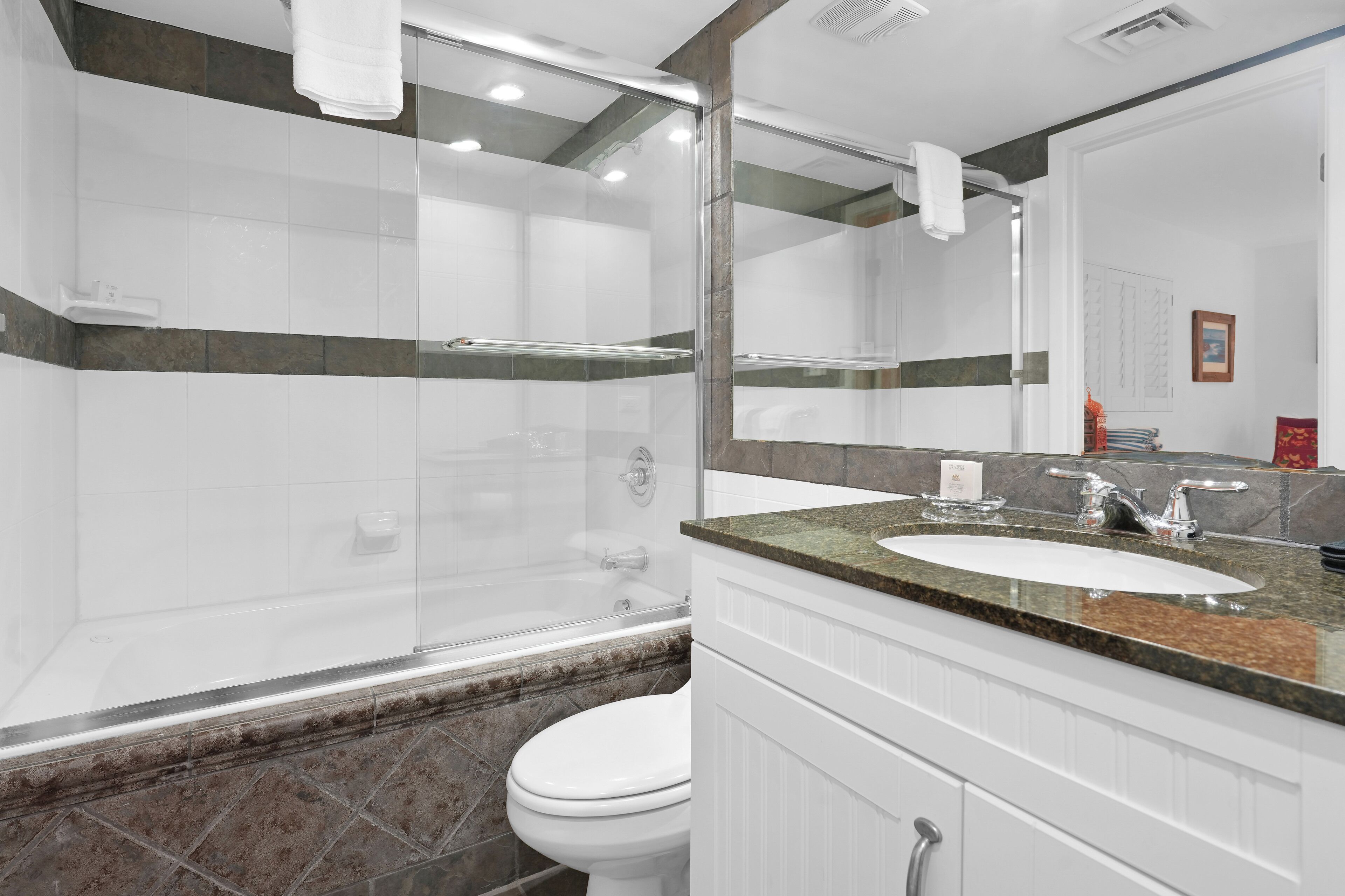 A modern bathroom with a glass-enclosed shower, a white toilet, a sink with a granite countertop, and a large mirror.