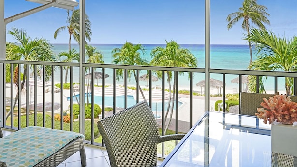 A balcony with a view of a beach, palm trees, and a pool. - Aqua Bay Club Condos (Seven Mile Beach)
