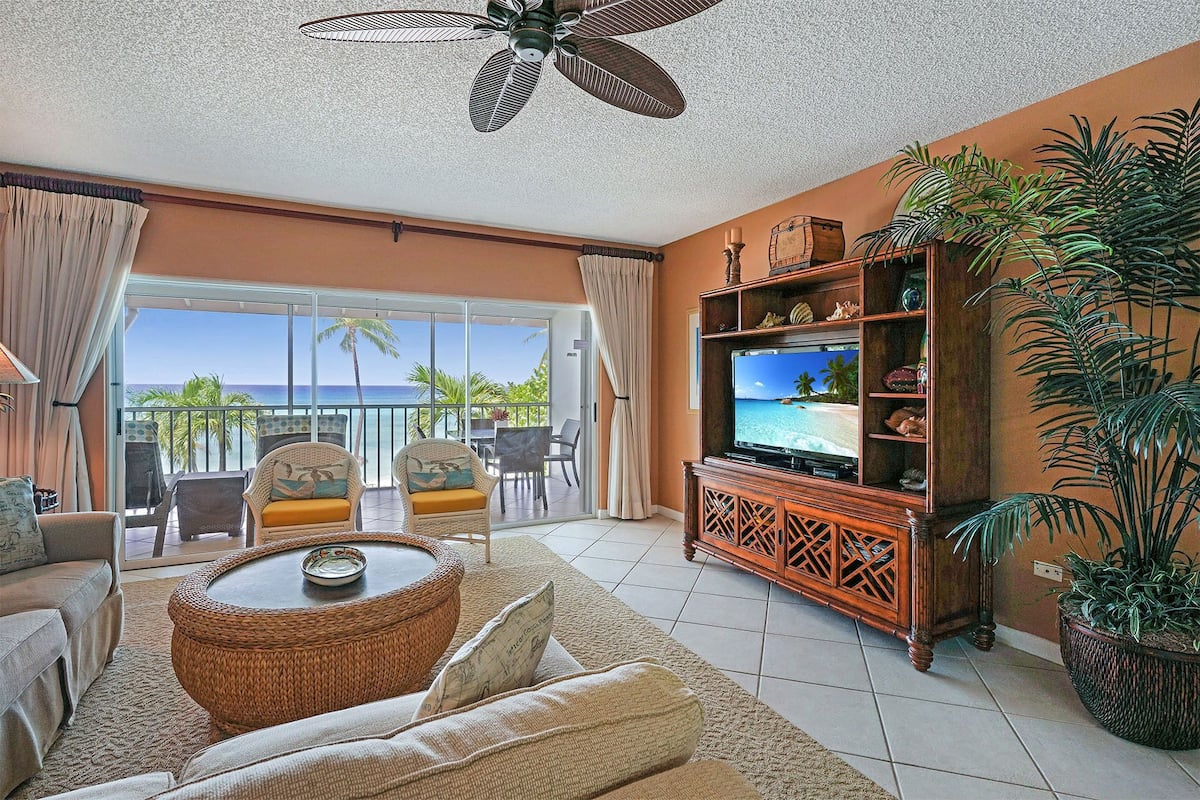 Una sala de estar con un televisor grande, muebles de mimbre y vista al océano.