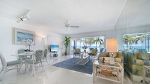 A spacious living room with a dining area, a ceiling fan, a large window with a view of palm trees, and a television. - Aqua Bay Club Condos (Seven Mile Beach)